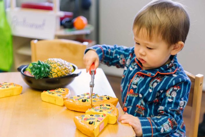 A young person is happily eating a snack at the table with a toddler in the background.