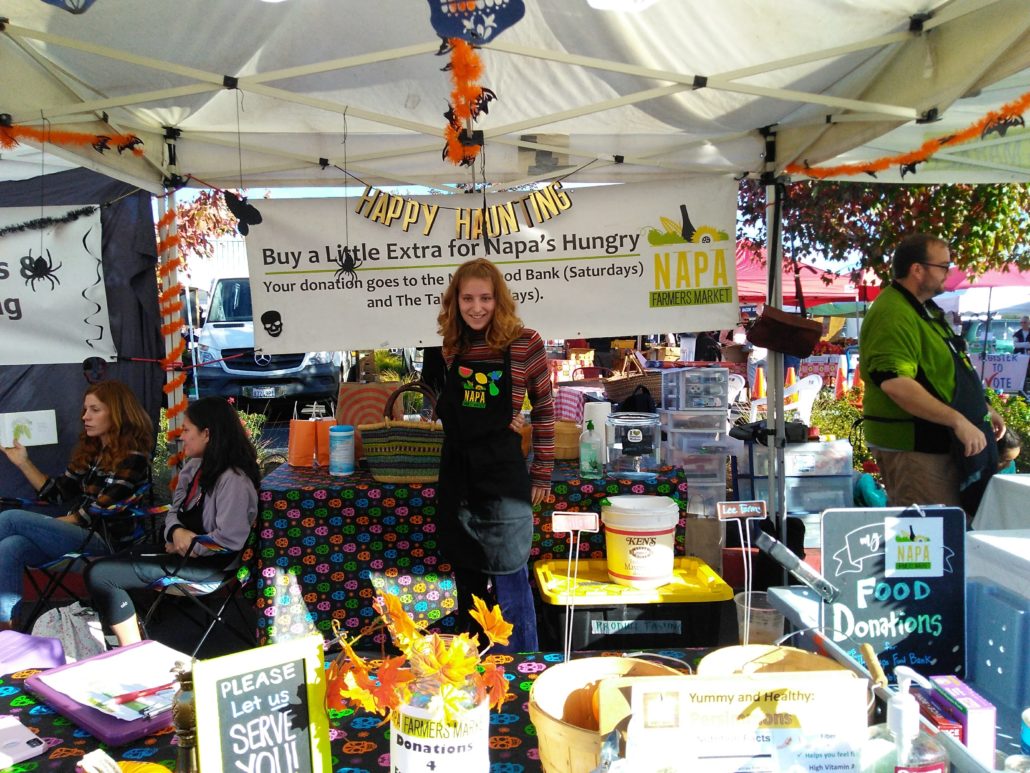 A person stands in an indoor marketplace, selling food to customers at a flea market retail shop with a text overhead that reads "Happy Haunting: Buy a Little Extra for Napa's Hungry".