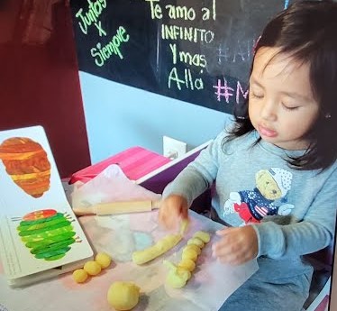 A toddler is smiling while holding a piece of child art with the text "Le Amo Al Infinito Y Mas Siempre Alla" written on it.