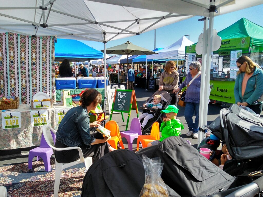 A person is selling their wares under a canopy in an outdoor marketplace, surrounded by other hawkers and people sitting in their shops in the street.