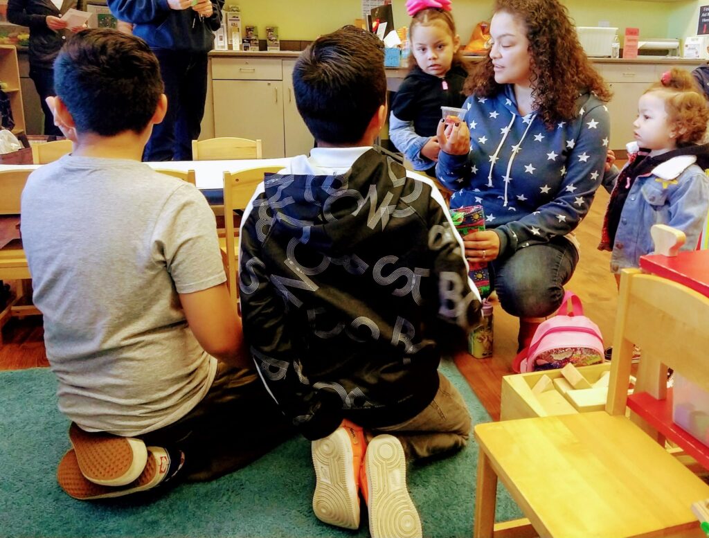 A group of children are learning together in a kindergarten classroom, seated around a table with furniture and human faces visible.